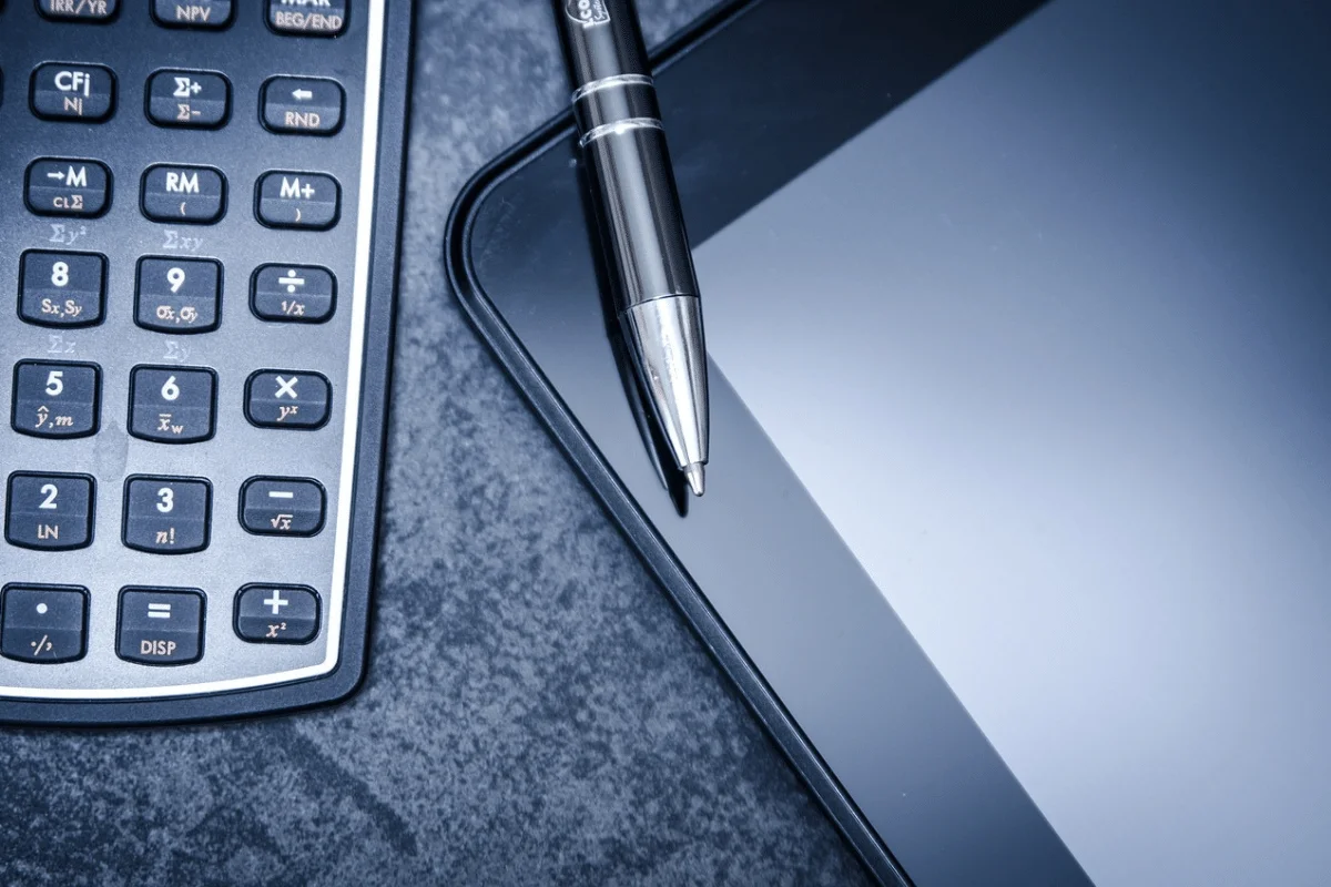 Close-up of a calculator, pen, and corner of a laptop on a desk.